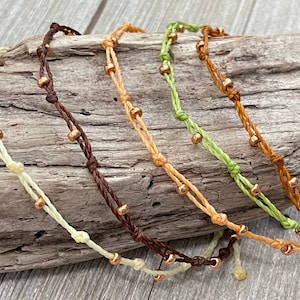 May include: Five bracelets made of thin, braided cord with small gold beads. The bracelets are in shades of brown, beige, and green.