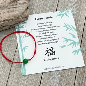 Puede incluir: Pulsera de cuerda roja con una cuenta de jade verde y el carácter chino para "bendición" escrito en negro. La tarjeta dice "Jade verde" y describe las propiedades y el significado de la piedra. La tarjeta también incluye el texto "Bendición-Fortuna" y el sitio web arieandco.etsy.com.