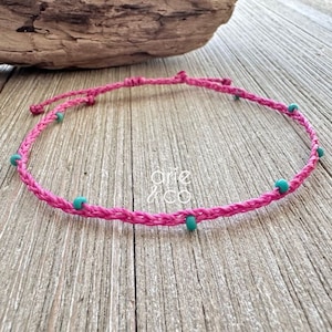 May include: A pink braided cord bracelet with turquoise beads. The bracelet is tied with a knot and has a simple design.