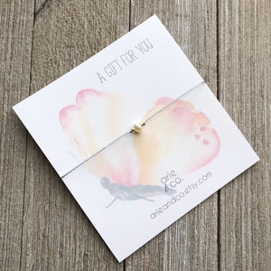 Puede incluir: Un delicado brazalete de cuerda gris con un solo dije de perla blanca. El brazalete se muestra en una tarjeta con un dise&ntilde;o de mariposa de acuarela y el texto "Un regalo para ti".