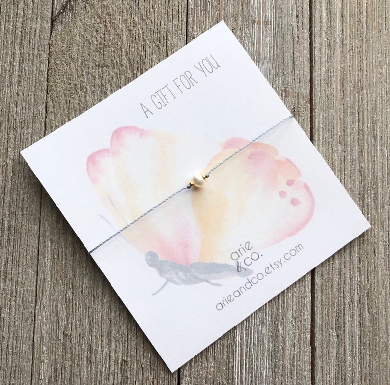 Puede incluir: Un delicado brazalete de cuerda gris con un solo dije de perla blanca. El brazalete se muestra en una tarjeta con un dise&ntilde;o de mariposa de acuarela y el texto "Un regalo para ti".