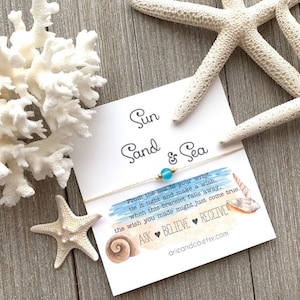 May include: A white card with the text "Sun Sand & Sea" and a blue and gold bracelet with a blue bead. The card has a watercolor illustration of seashells and the text "From the sea to your wrist, tie it tight and make a wish, when this bracelet falls away, the wish you made might just come true. ASK BELIEVE RECEIVE arieandco.etsy.com"