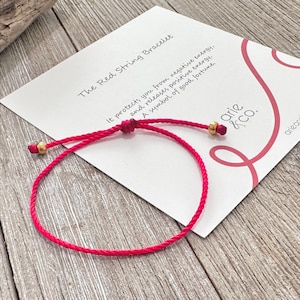 May include: A thin red string bracelet featuring two small gold-toned metallic beads and a sliding knot closure. The bracelet is displayed on a white card with the text The Red String Bracelet, arie & co., and descriptive text about energy.