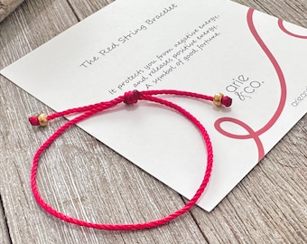 May include: A thin red string bracelet featuring two small gold-toned metallic beads and a sliding knot closure. The bracelet is displayed on a white card with the text The Red String Bracelet, arie & co., and descriptive text about energy.
