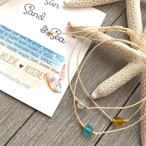 May include: Three white string bracelets with different colored glass beads. The beads are blue, yellow, and white. The bracelets are on a wooden surface with a starfish and a card that says "Sun Sand & Sea" and has a watercolor beach scene.