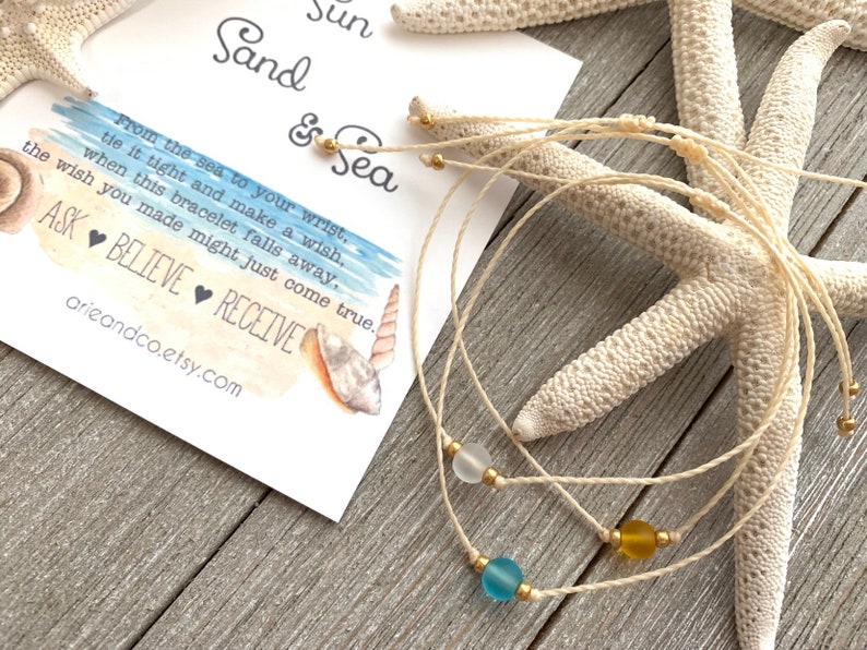 May include: Three white string bracelets with different colored glass beads. The beads are blue, yellow, and white. The bracelets are on a wooden surface with a starfish and a card that says "Sun Sand & Sea" and has a watercolor beach scene.