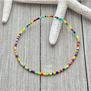 May include: A colorful beaded bracelet with a rainbow of colors, including red, orange, yellow, green, blue, and purple. The bracelet is made of small, round beads and is strung on a thin, clear elastic cord.