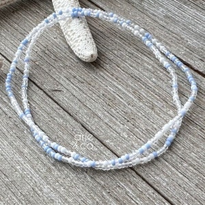 May include: A delicate double strand necklace made with tiny white and light blue beads. The necklace is layered and has a subtle ombre effect.