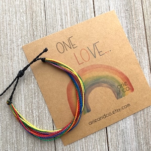May include: A rainbow colored friendship bracelet with black cord ties. The bracelet is on a brown card with the words "ONE LOVE..." and a rainbow painted on it. The card also has the text "arie & co." and "arieandco.etsy.com".