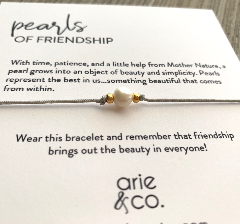 May include: A gray string bracelet with a white pearl and gold beads. The card reads "pearls of friendship" and includes a quote about the beauty of friendship. The card is signed "arie & co."