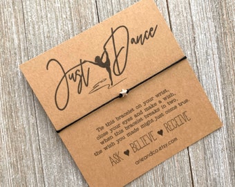 May include: A brown card with the text "Just Dance" in black script. A black string bracelet with a silver star charm is tied around the card. The card also includes the text "Tie this bracelet on your wrist, close your eyes and make a wish, when this bracelet breaks in two, the wish you made might just come true. ASK BELIEVE RECEIVE arieandco.etsy.com"
