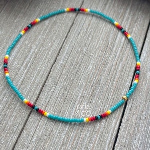 May include: A turquoise beaded necklace with red, yellow, white, and black accents. The necklace has a gold bead at the end.
