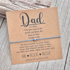 May include: A brown card with the word "Dad" in large, stylized lettering. The card defines "Dad" as "the best man in the world, our hero, the man we love. Irreplaceable."  A blue string bracelet with a silver star charm is tied around the card. The card also includes the text "Tie this bracelet on your wrist, close your eyes and make a wish, when this bracelet breaks in two, the wish you made might just come true. Ask Believe Receive arieandco.etsy.com"
