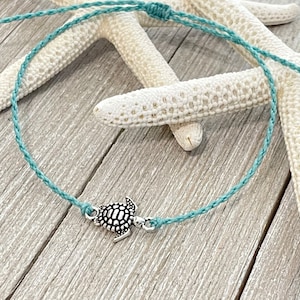 May include: A turquoise blue braided cord bracelet with a silver turtle charm. The bracelet is displayed on a white starfish.
