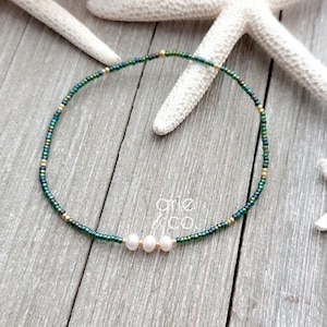May include: A delicate green and gold beaded necklace with three white pearls. The necklace is on a wooden surface.