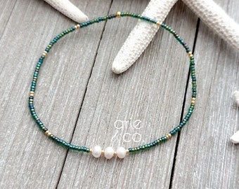 May include: A delicate green and gold beaded necklace with three white pearls. The necklace is on a wooden surface.