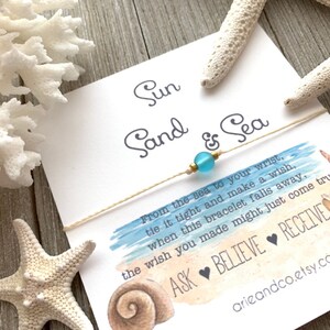 May include: A white card with blue watercolor background and the text "Sun Sand & Sea" with a string bracelet with a light blue bead. The text on the card reads "From the sea to your wrist, tie it tight and make a wish, when this bracelet falls away, the wish you made might just come true. ASK BELIEVE RECEIVE arieandco.etsy.com"