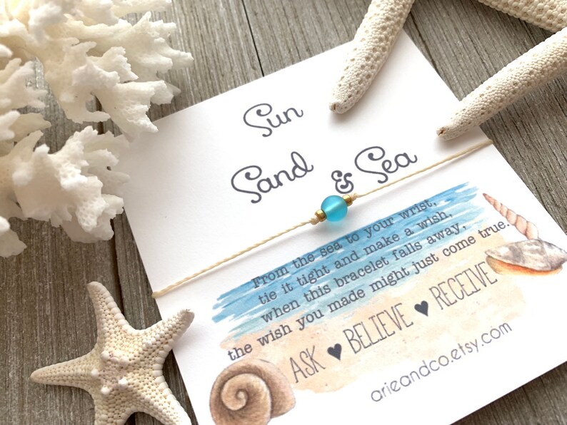 May include: A white card with blue watercolor background and the text "Sun Sand & Sea" with a string bracelet with a light blue bead. The text on the card reads "From the sea to your wrist, tie it tight and make a wish, when this bracelet falls away, the wish you made might just come true. ASK BELIEVE RECEIVE arieandco.etsy.com"