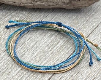 May include: A set of four friendship bracelets in shades of blue, green, and beige. The bracelets are made of thin, braided cord and are tied with a simple knot.