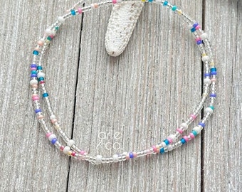 May include: A delicate double-strand bracelet with tiny white beads and colorful accents. The bracelet features a mix of pink, blue, yellow, and green beads.