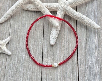 May include: A red beaded bracelet with a single white pearl accent. The bracelet is on a wooden surface with a starfish in the background.