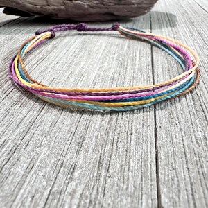 May include: A multi-strand cord bracelet featuring thin, twisted threads in shades of purple, yellow, teal, and tan. The bracelet is secured with a dark purple knotted closure, creating a layered appearance with a circular shape and smooth texture.