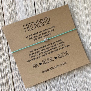 May include: A light blue friendship bracelet with a silver flower charm. The card reads "Friendship is not what we have in life, but who we have in our life that matters. Tie this bracelet on your wrist, close your eyes and make a wish, when this bracelet breaks in two, the wish you made might just come true. Ask Believe Receive arieandco.etsy.com"