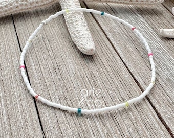 May include: White beaded necklace with colorful accents. The necklace is made of small white beads with a few colorful beads interspersed throughout. The necklace is lying on a wooden surface.