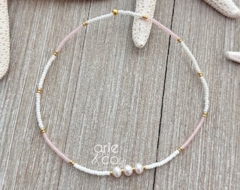 May include: A delicate white and pink beaded necklace with three small pearl accents. The necklace is made with tiny seed beads and has a gold clasp.