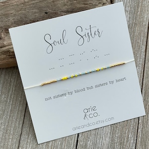 May include: A white card with the text "Soul Sister" written in black cursive. Below the text is a white string with a beaded bracelet made of gold, silver, and yellow beads. The card also includes the text "not sisters by blood but sisters by heart" and the company name "arie & CO." with the website address "arieandco.etsy.com".