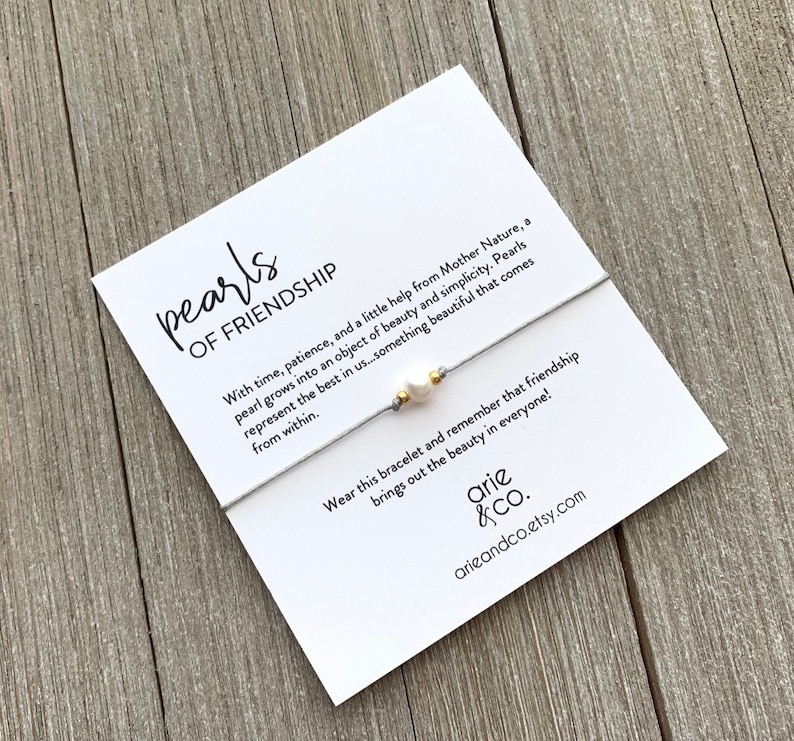 May include: A white card with the text "pearls of friendship" and a gray string bracelet with a white pearl and gold bead charm. The card reads "With time, patience, and a little help from Mother Nature, a pearl grows into an object of beauty and simplicity. Pearls represent the best in us...something beautiful that comes from within. Wear this bracelet and remember that friendship brings out the beauty in everyone!" and is signed "arie &Co. arieandco.etsy.com"