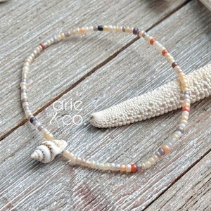May include: A delicate bracelet made with small, colorful beads and a single seashell charm. The bracelet is displayed on a wooden surface with a starfish.