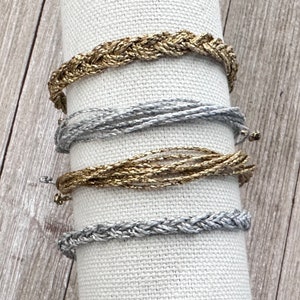 May include: Four bracelets made of braided silver and gold thread. The bracelets are displayed on a white fabric background.