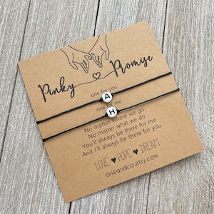 May include: Two black string bracelets with white letter beads spelling "AH". The bracelets are on a card with the text "Pinky Promise" and a poem about friendship.