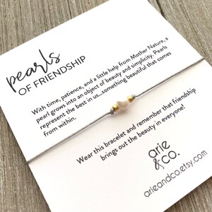 May include: A gray string bracelet with a white pearl charm and gold beads. The card reads "pearls of friendship" and includes a quote about the beauty of pearls and friendship.