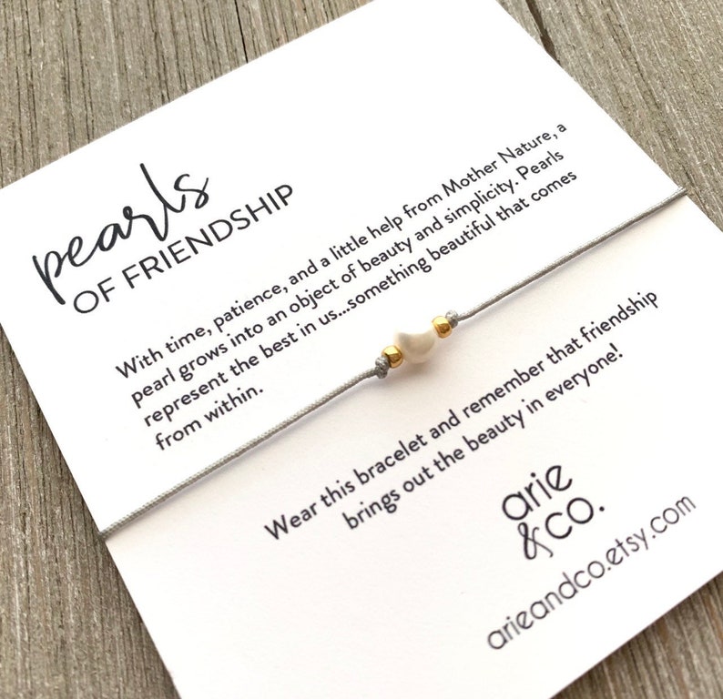 May include: A gray string bracelet with a white pearl charm and gold beads. The card reads "pearls of friendship" and includes a quote about the beauty of pearls and friendship.