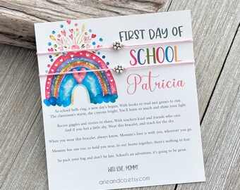 May include: A pink and white card with a rainbow design and the text "First Day of School Patricia". Two pink friendship bracelets with silver beads are attached to the card.