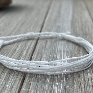 May include: A white braided cord bracelet, made of multiple strands of thin white cord, on a wooden surface.