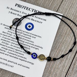 May include: A black string bracelet featuring seven knots, a blue and white evil eye bead, a gold-toned St. Benedict medal, and a black bead. The bracelet is displayed on a white card with the text PROTECTION, ST. BENEDICT, EVIL EYE & AZABACHE.