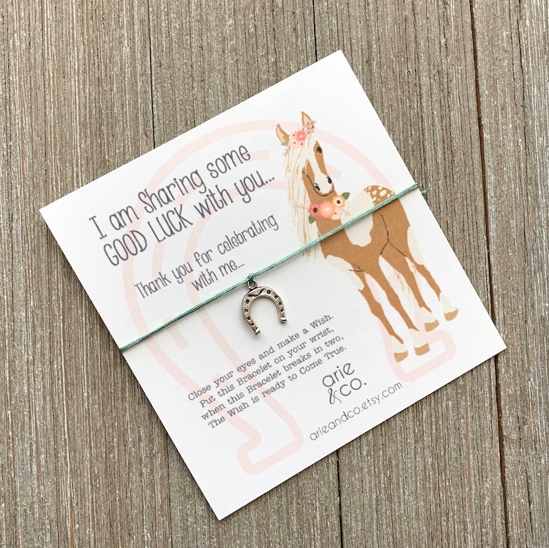 Op de afbeelding: Een witte kaart met een roze en witte paardenillustratie en de tekst "I am Sharing some GOOD LUCK with you... Thank you for celebrating with me... Close your eyes and make a Wish. Put this Bracelet on your wrist, when this Bracelet breaks in two, The Wish is ready to Come True. arie arieandco.etsy.com". Een turquoise touwtjesarmband met een zilveren hoefijzerbedel is aan de kaart bevestigd.