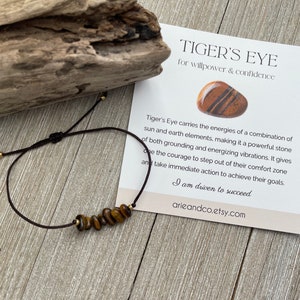 May include: A brown string bracelet with gold beads and tiger's eye stones. The bracelet is tied with a knot and has a small gold bead at the end. The bracelet is accompanied by a card that reads "Tiger's Eye for willpower & confidence." The card also includes a description of the stone's properties and the phrase "I am driven to succeed." The card is on a wooden surface.
