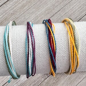 May include: Four braided bracelets in different color combinations. The bracelets are made of thin, woven cords in shades of green, blue, white, red, and yellow.