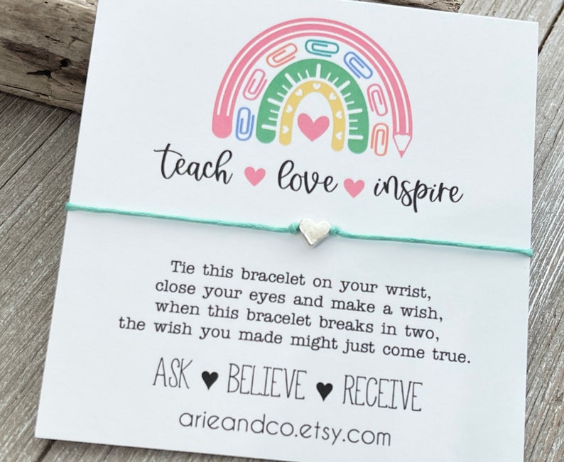 May include: A white card with a rainbow made of colourful paper clips and the words "teach love inspire" in black script. The card also includes the text "Tie this bracelet on your wrist, close your eyes and make a wish, when this bracelet breaks in two, the wish you made might just come true. ASK BELIEVE RECEIVE arieandco.etsy.com".