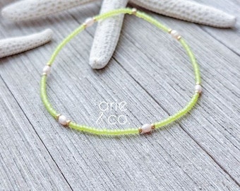 May include: A delicate green and white beaded bracelet with small pearl accents. The bracelet is on a wooden surface.