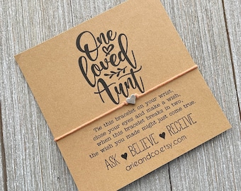 May include: A brown card with the text "One loved Aunt" in black script. A pink string bracelet with a silver heart charm is attached to the card. The text on the card reads: "Tie this bracelet on your wrist, close your eyes and make a wish, when this bracelet breaks in two, the wish you made might just come true. ASK BELIEVE RECEIVE arieandco.etsy.com"
