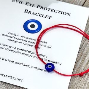May include: A red string bracelet featuring a small, round, blue evil eye bead with a white and black center. The bracelet is displayed on a white card printed with the text Evil Eye Protection Bracelet and a description of the item.
