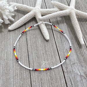 May include: A white beaded necklace with red, yellow, and black accents. The necklace is on a wooden surface with two white starfish.
