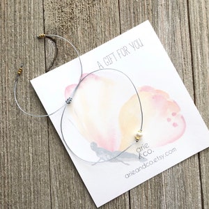 Puede incluir: Un brazalete de cuerda gris con cuentas doradas y un dije de perla blanca. El brazalete est&aacute; sobre una tarjeta con un dise&ntilde;o de mariposa acuarela y el texto "Un regalo para ti". La tarjeta tambi&eacute;n tiene el texto "arie & co." y "arieandco.etsy.com".