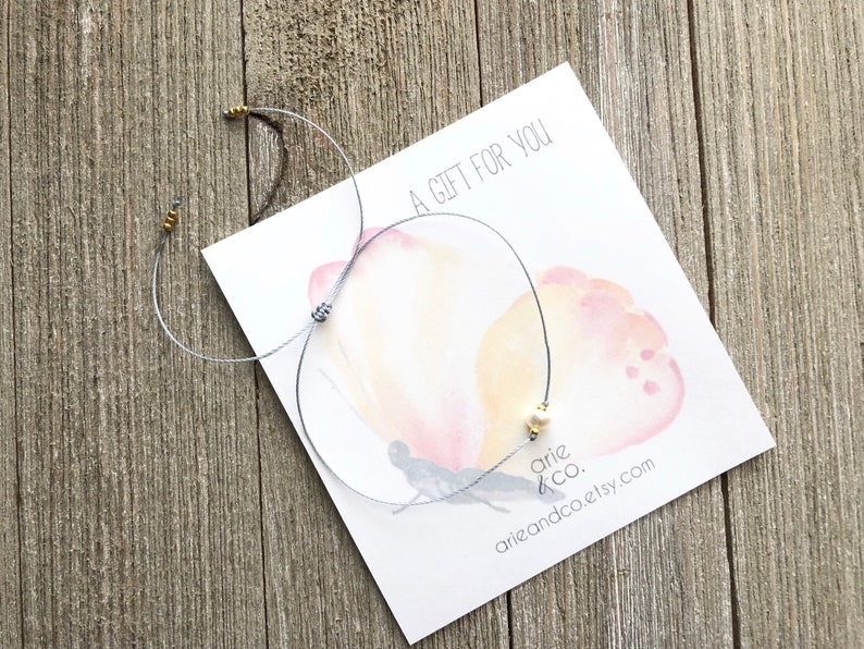 Puede incluir: Un brazalete de cuerda gris con cuentas doradas y un dije de perla blanca. El brazalete est&aacute; sobre una tarjeta con un dise&ntilde;o de mariposa acuarela y el texto "Un regalo para ti". La tarjeta tambi&eacute;n tiene el texto "arie & co." y "arieandco.etsy.com".