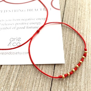 May include: Red string bracelet with gold beads. The card reads "The Red String Bracelet. It protects you from negative energy and releases positive energy. A symbol of good fortune. arie & co arieandco.etsy.com"